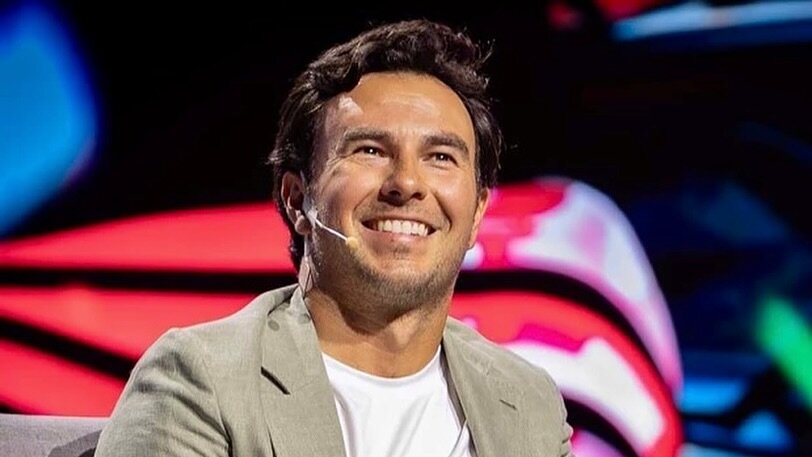 Sergio "Checo" Pérez sonriente, con micrófono en mano, participando en un evento público, con estructura metálica de escenario al fondo.