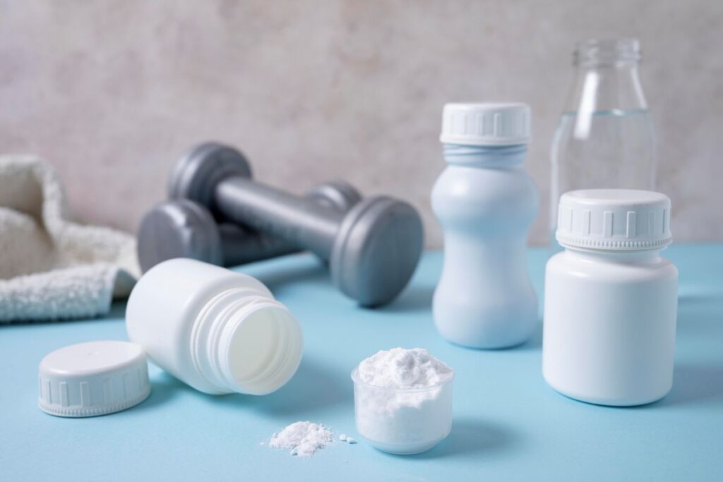 Suplementos en botellas y polvo blanco en un vaso medidor junto a pesas y una botella de agua, representando la suplementación con creatina para mejorar el rendimiento físico y la salud muscular.Suplementos en botellas y polvo blanco en un vaso medidor junto a pesas y una botella de agua, representando la suplementación con creatina para mejorar el rendimiento físico y la salud muscular.