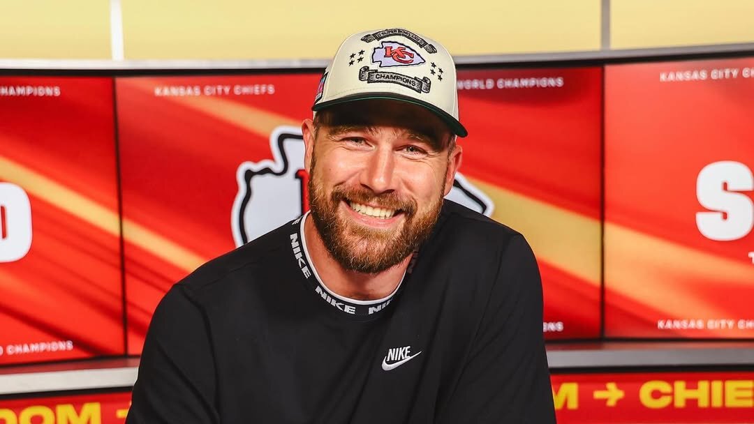 Travis Kelce, futuro esposo de Taylor Swif, vestido con camiseta negra Nike y gorra de los Kansas City Chiefs, firma documentos en una mesa frente a pantallas con el logo del equipo, destacando su papel como estrella de la NFL. 