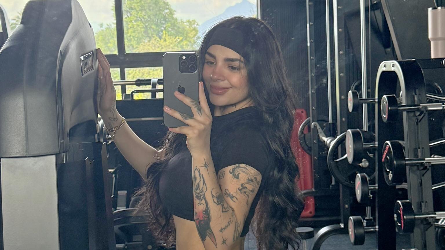 Foto de Karely Ruiz en el gimnasio, tomándose una selfie frente al espejo. Lleva una camiseta negra corta y leggings color gris claro, con el cabello suelto y una cinta negra, mostrando su figura atlética y tatuajes en el brazo.