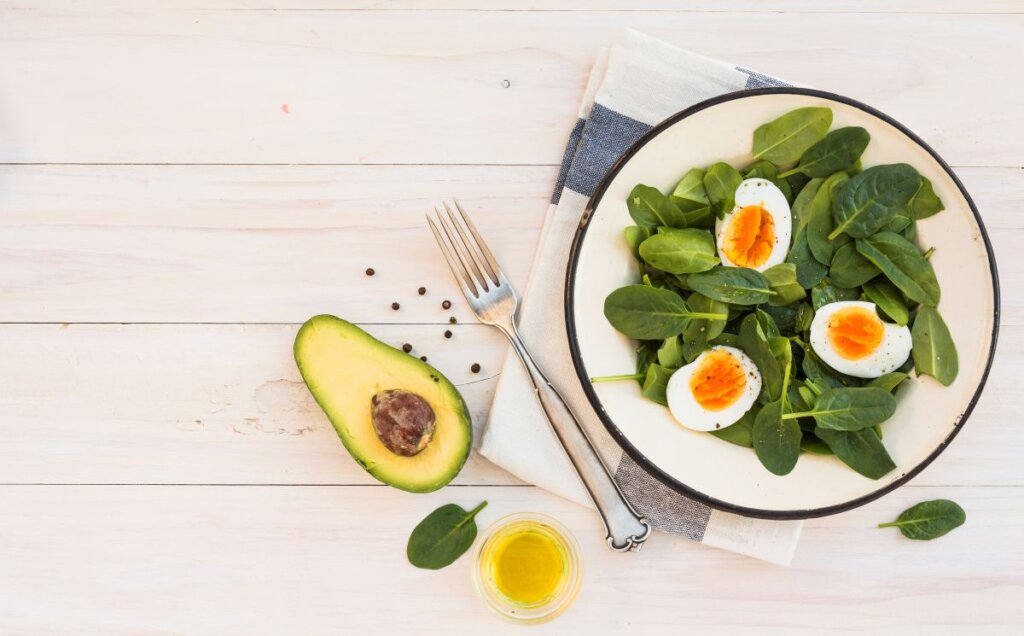 Plato de espinacas frescas con huevos cocidos y aguacate, acompañado de aceite de oliva, alimentos característicos de la dieta cetogénica por su alto contenido en grasas saludables y bajo aporte de carbohidratos.